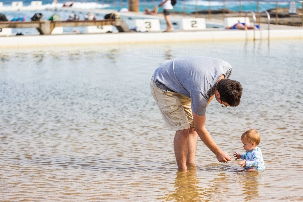 Happy young baby girl sitting in shallow sea water in ocean pool at beach in summer - Australian Stock Image