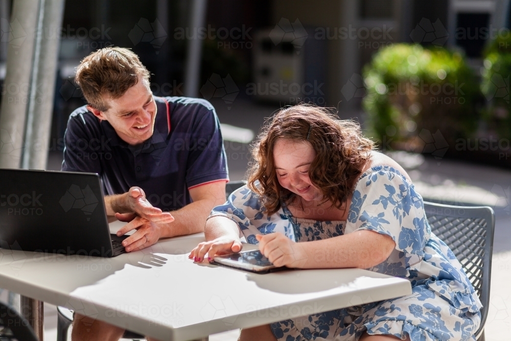 Happy young disability worker and his client with down syndrome working on devices at table - Australian Stock Image