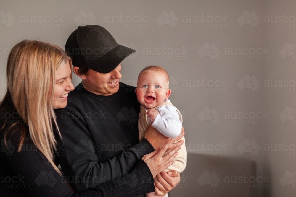 Happy young family smiling together in home dad holding baby boy with big smile - Australian Stock Image