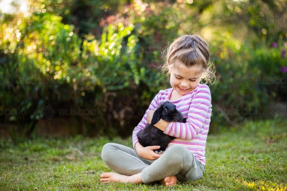 Happy young girl cuddling black pet bunny rabbit in garden outside - Australian Stock Image