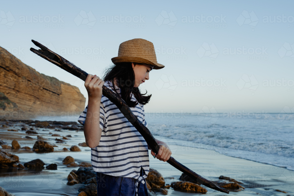 Happy young girl wearing a sunhat holding a stick having fun playing at the beach at sunset - Australian Stock Image