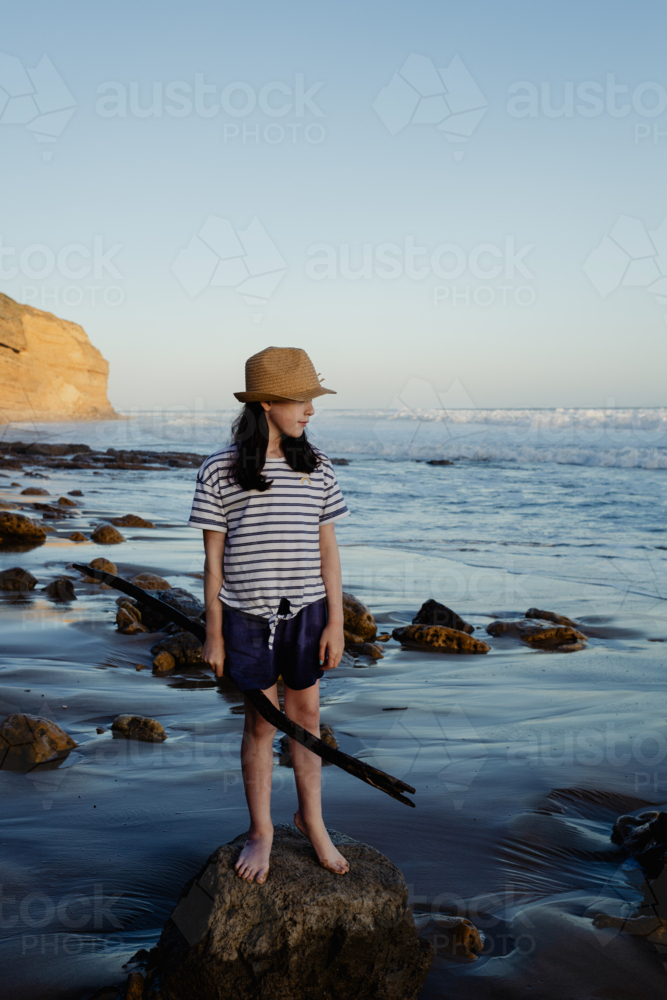 Happy young girl wearing a sunhat on a rock holding a stick, having fun playing at the beach - Australian Stock Image