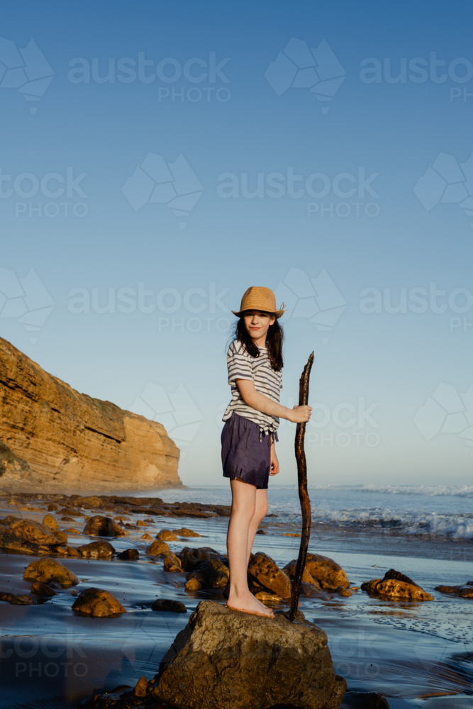 Happy young girl wearing a sunhat standing on a rock holding a stick playing at the beach - Australian Stock Image