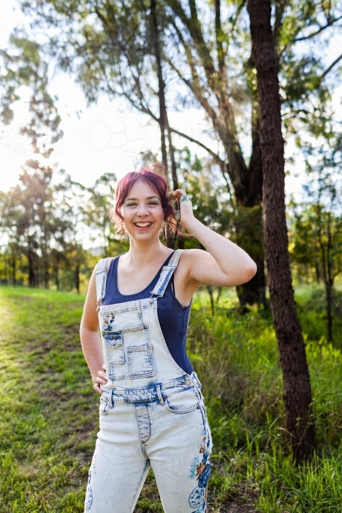 Happy young lady artist with paintbrush tucking hair behind ear - Australian Stock Image