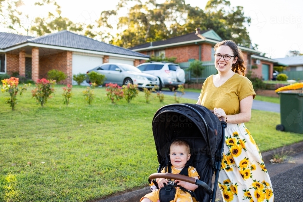 Happy young mother pushing baby in pram down street in front of houses - Australian Stock Image