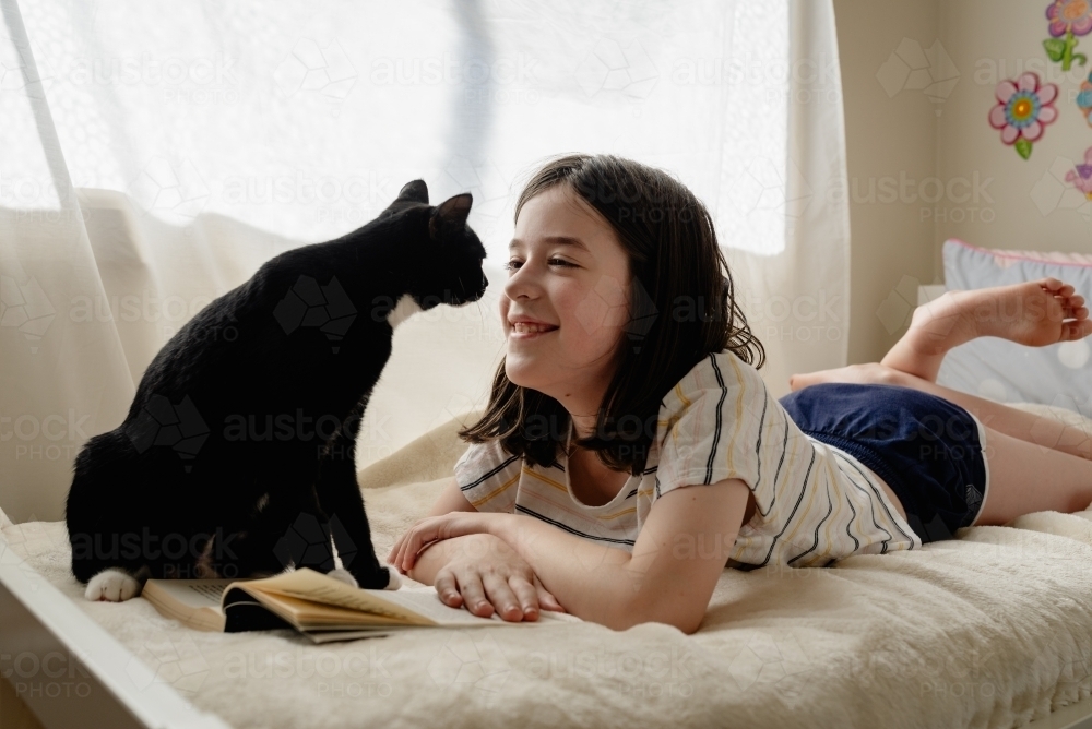 Happy young preteen girl in her bedroom lying on her bed with her cat while she reads a book - Australian Stock Image