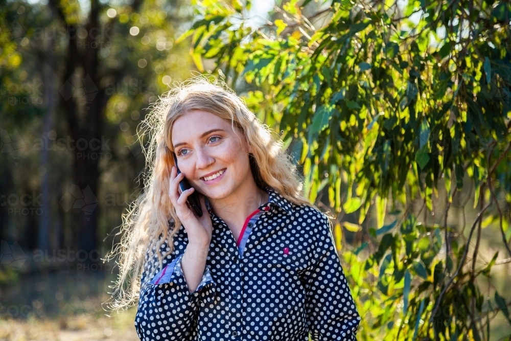 Happy young woman outside under shady tree talking on her mobile phone - Australian Stock Image