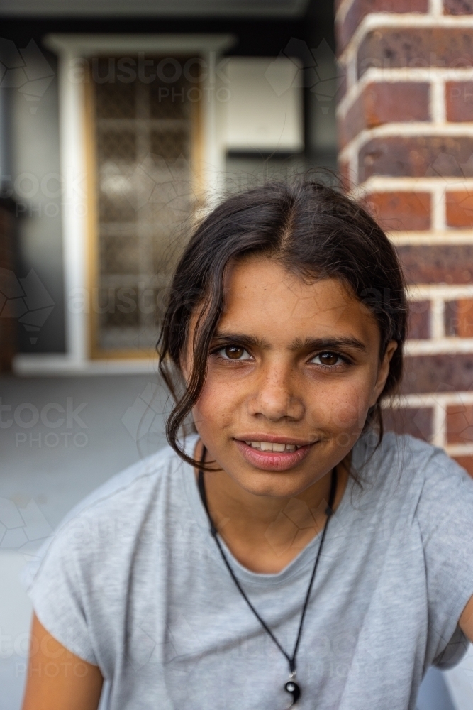 head and shoulders of aboriginal girl looking at camera with brick wall and door behind - Australian Stock Image