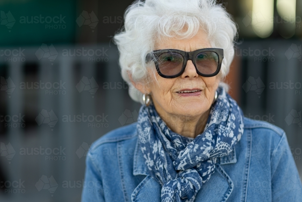 head and shoulders of elderly white-haired lady wearing denim with sunglasses and a scarf - Australian Stock Image