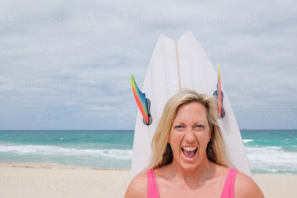 Head shot of Woman in bathers looking at camera on beach holding surfboard behind head - Australian Stock Image