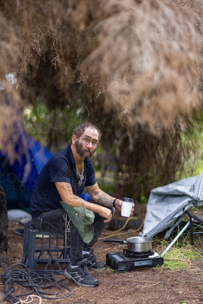 Homeless man sitting on crate outside his tent with a coffee - Australian Stock Image