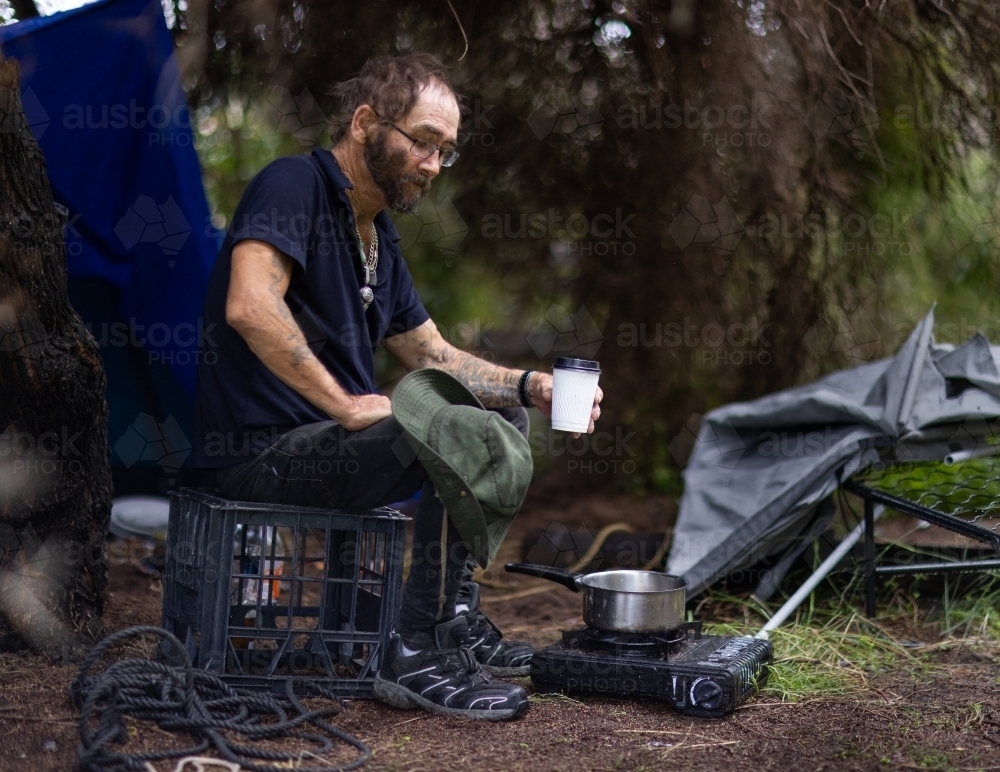 homeless man sitting on milk crate near butane stove in front of tarpaulin camp - Australian Stock Image