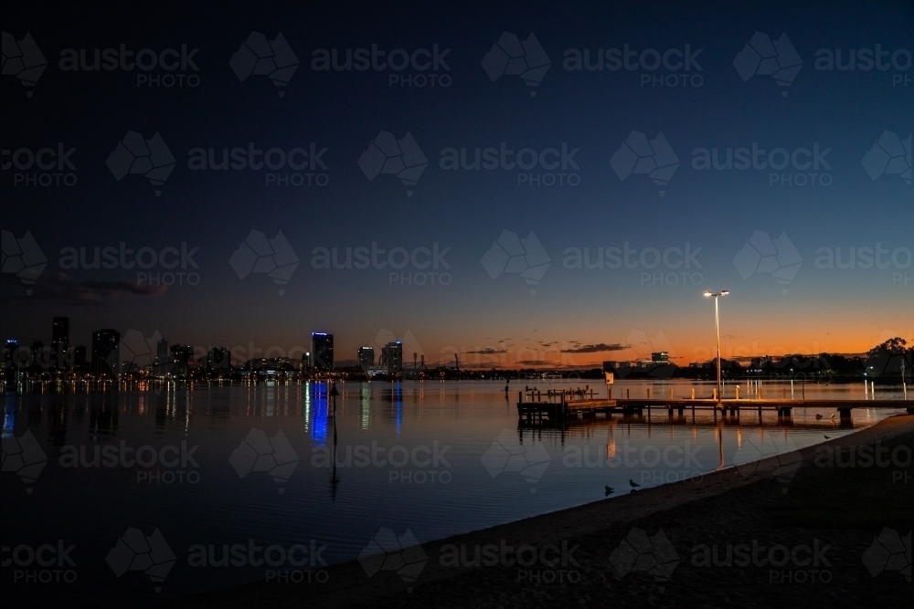 horizontal shot of a jetty with lamp post during sunset with buildings reflected by the water - Australian Stock Image