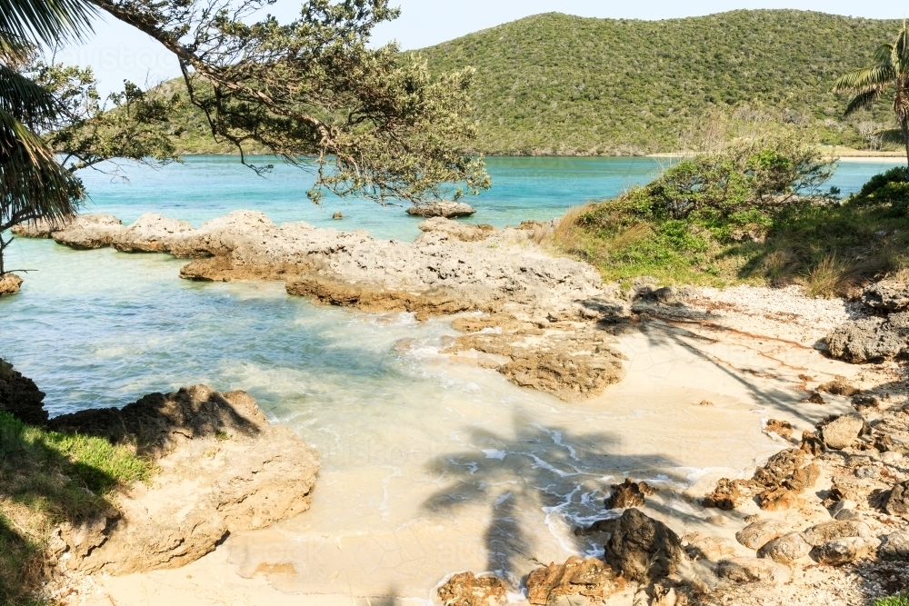 Horizontal shot of a remote beach with rocks mountains and trees on a sunny day with clear skies - Australian Stock Image