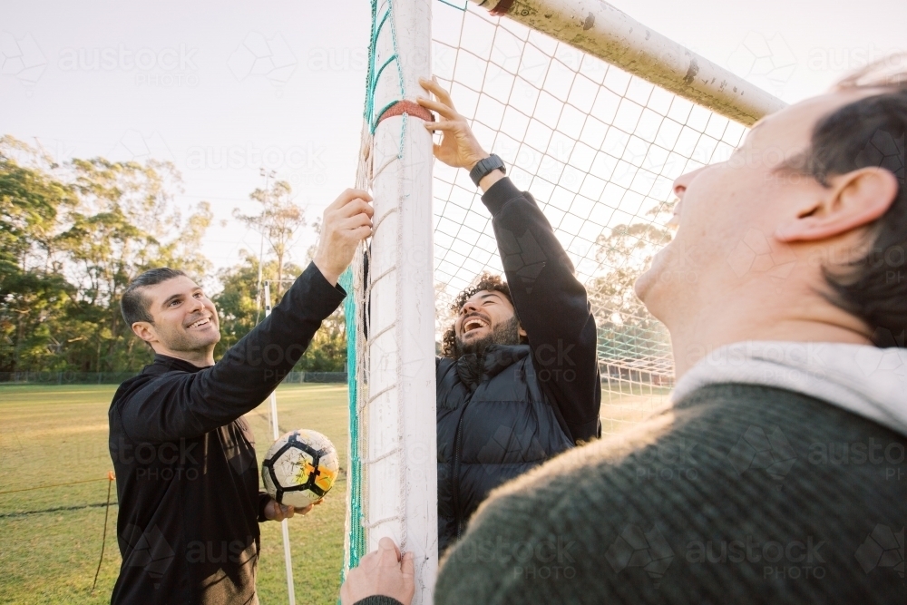 horizontal shot of three men smiling one holding a soccer ball and two touching the net - Australian Stock Image