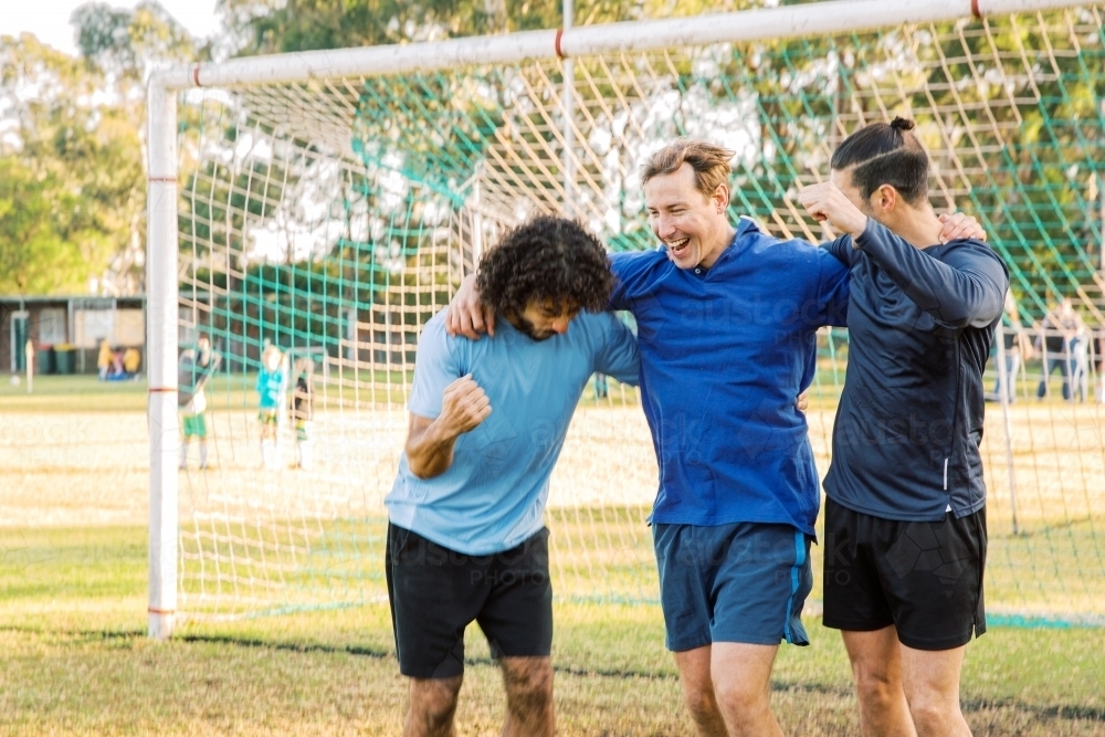 horizontal shot of three men smiling with arms around their shoulder with a goal in the background - Australian Stock Image