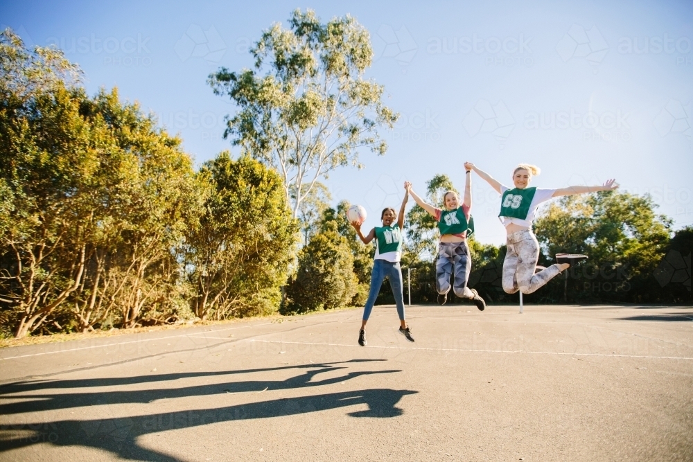 horizontal shot of three young women jumping in mid air on a sunny day with trees in the background - Australian Stock Image