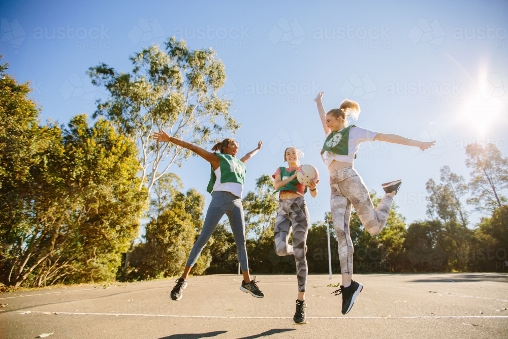 horizontal shot of three young women jumping in mid air with trees at the back on a sunny day - Australian Stock Image