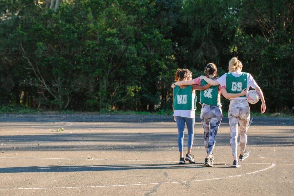 horizontal shot of three young women walking away with arms wrapped around each other on a sunny day - Australian Stock Image