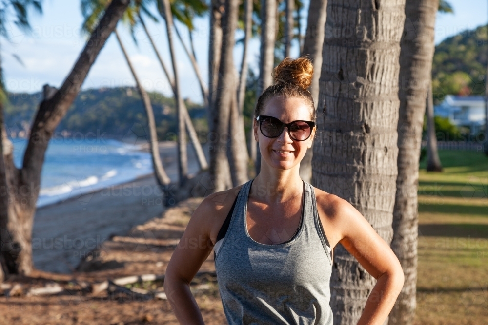 horizontal shot of woman smiling with sunglasses on by the beach with trees, mountains and waves - Australian Stock Image