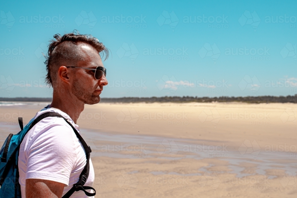 horizontal side view shot of a man wearing white shirt with backpack on the seashore - Australian Stock Image