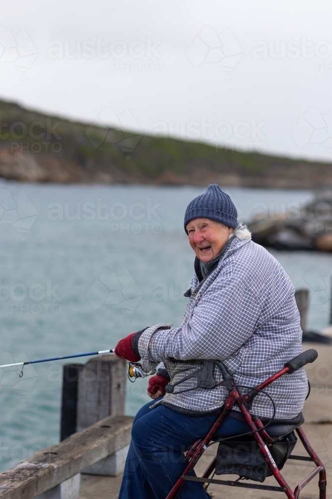 independent elderly lady sitting on wheely walker fishing rugged up in cold weather - Australian Stock Image