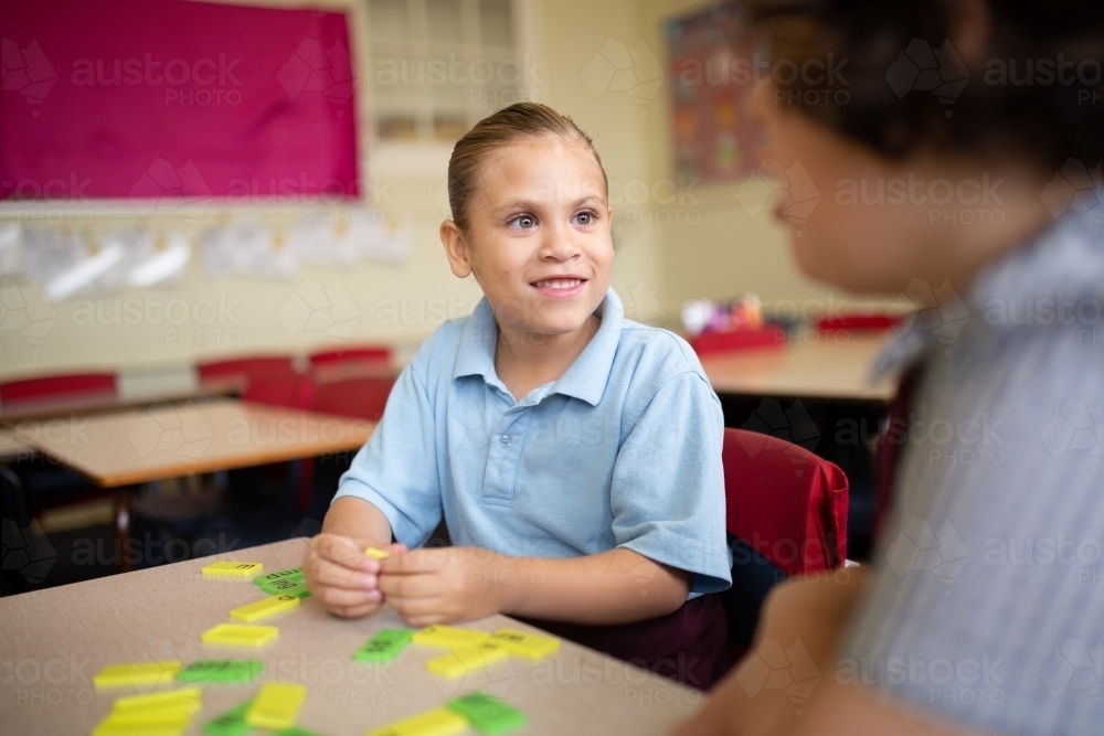 Indigenous girl primary school student sitting smiling at her friend working with word tiles - Australian Stock Image