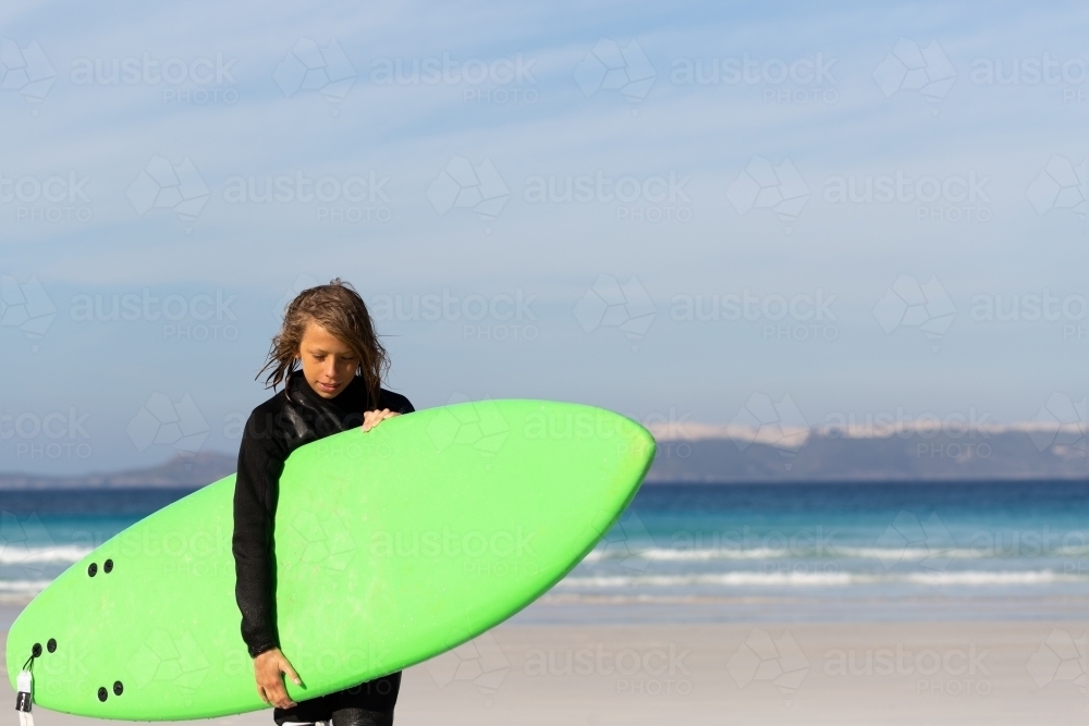 Kid in wetsuit carrying green surfboard at he beach - Australian Stock Image