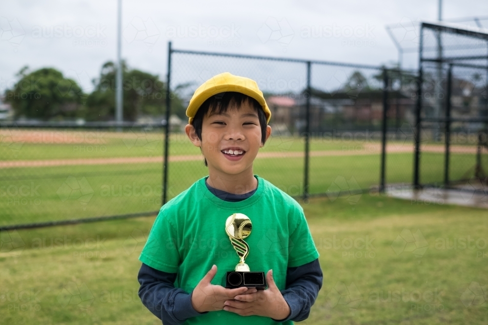 Kid smiling with a trophy, standing in front of a baseball diamond - Australian Stock Image