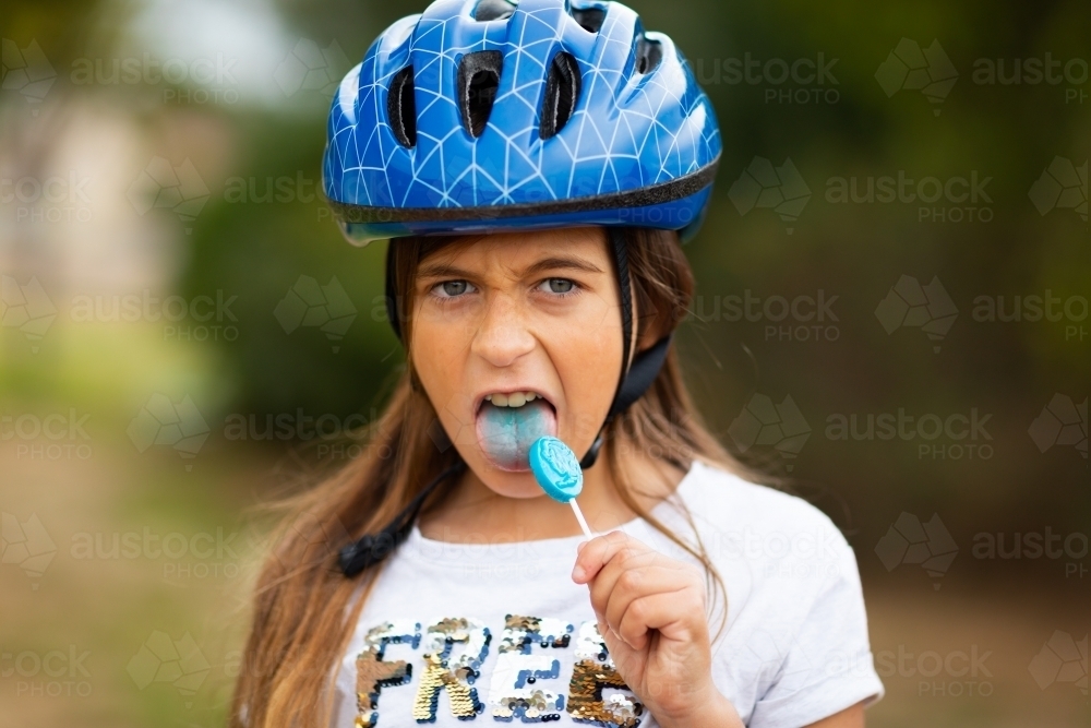 Kid with blue tongue and blue lollypop wearing blue bike helmet - Australian Stock Image