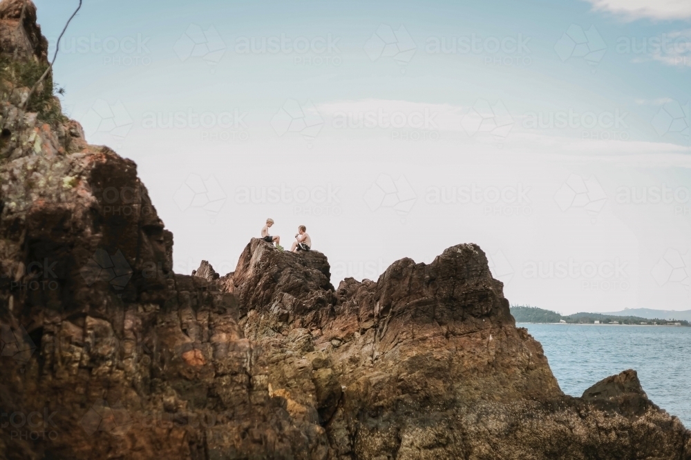 Kids sitting on rocks by the beach at Coral Beach near Shute Harbour in the Whitsundays - Australian Stock Image