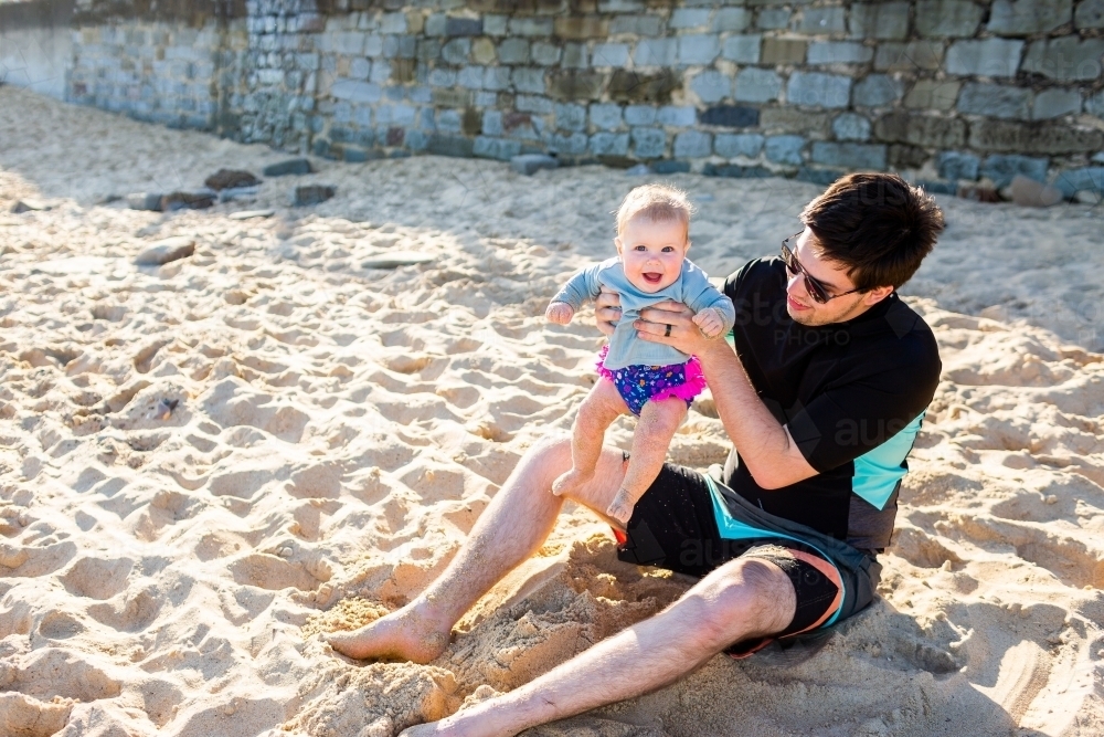 laughing baby with father playing in sand at beach - Australian Stock Image