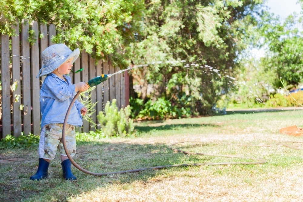 Little boy in blue hat playing with hose and water outside on hot summer day - Australian Stock Image