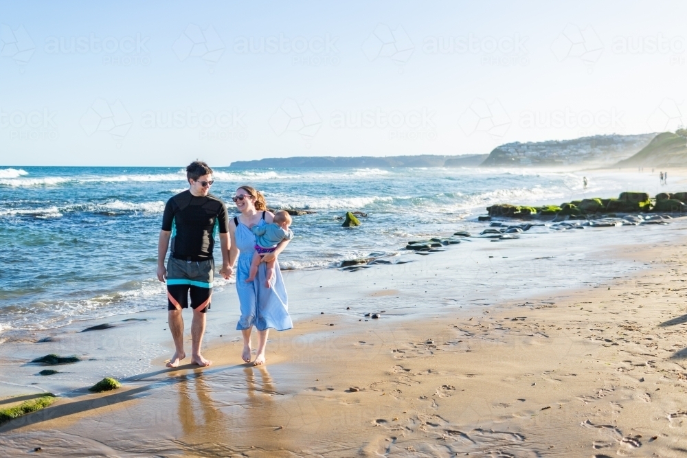 Little family walking down beach in afternoon light together a couple with baby - Australian Stock Image