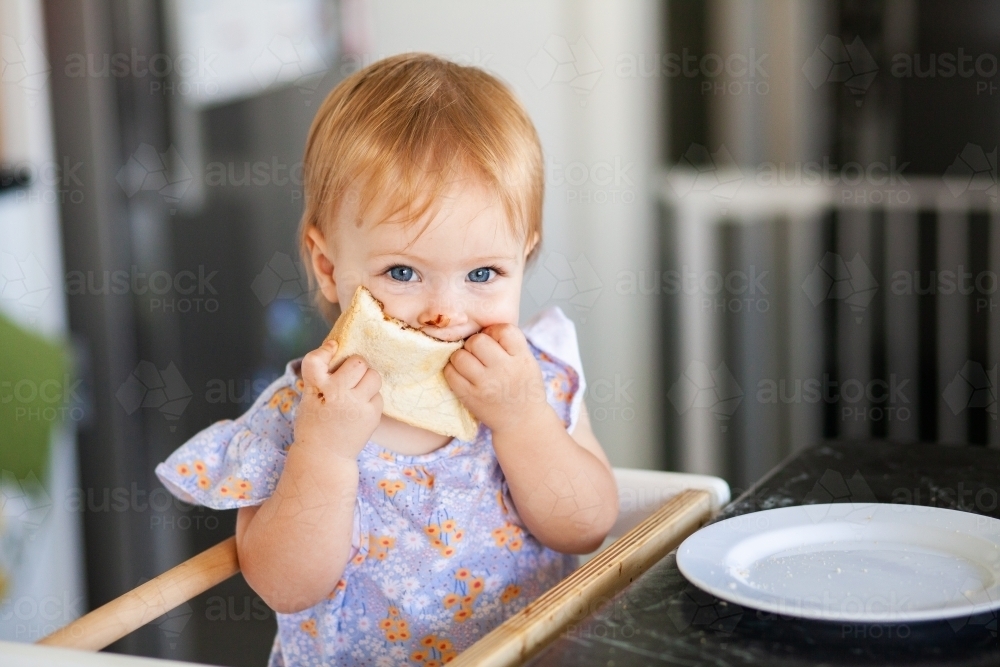 Little girl eating vegemite toast standing beside kitchen bench - Australian Stock Image