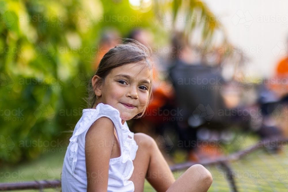little girl smiling looking over shoulder, with adults blurred in background - Australian Stock Image