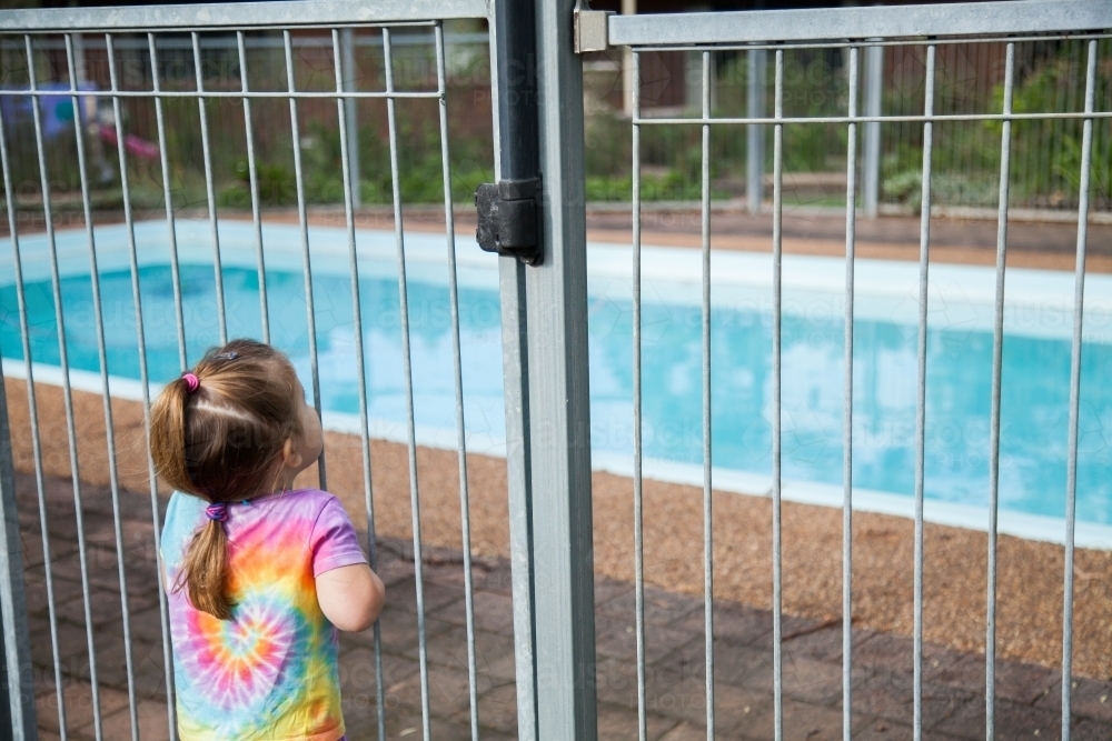 Little girl standing safely outside pool fence gate looking in - Australian Stock Image