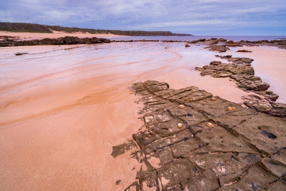 Long angled view of creek water flowing over a wide sandy beach around rocky edges at twilight - Australian Stock Image