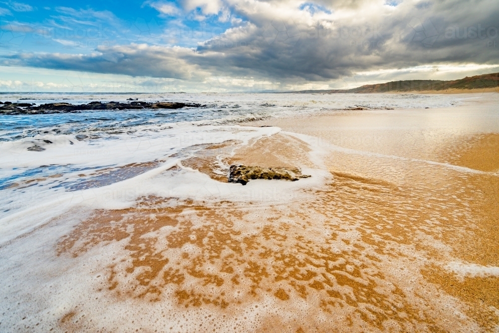 Long angled view of frothing waves running up a golden sandy beach around a rock - Australian Stock Image