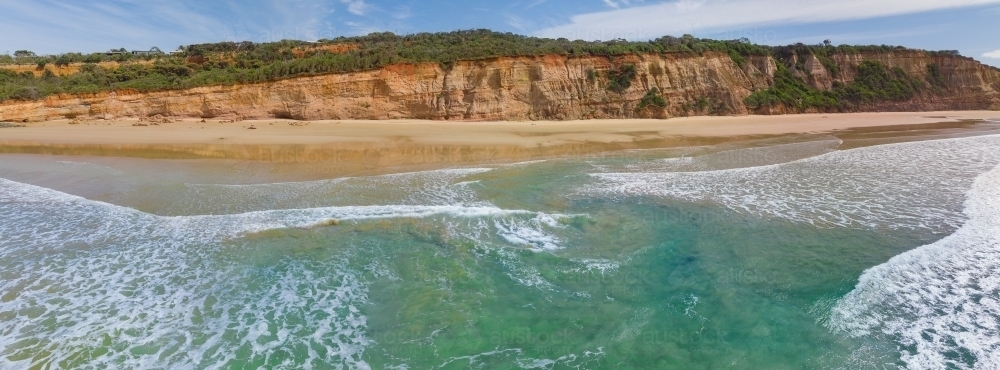 Looking inland over waves crashing onto a beach under rugged cliffs - Australian Stock Image