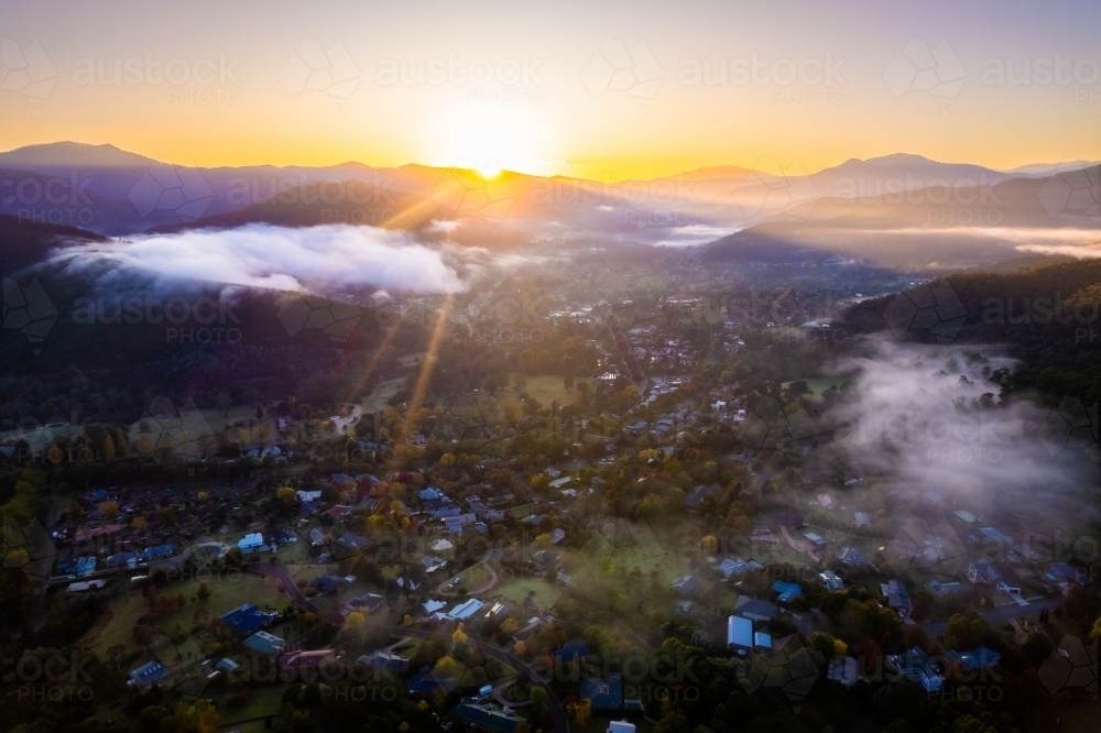 Looking over an autumn filled town through clouds and morning sunrise rays - Australian Stock Image