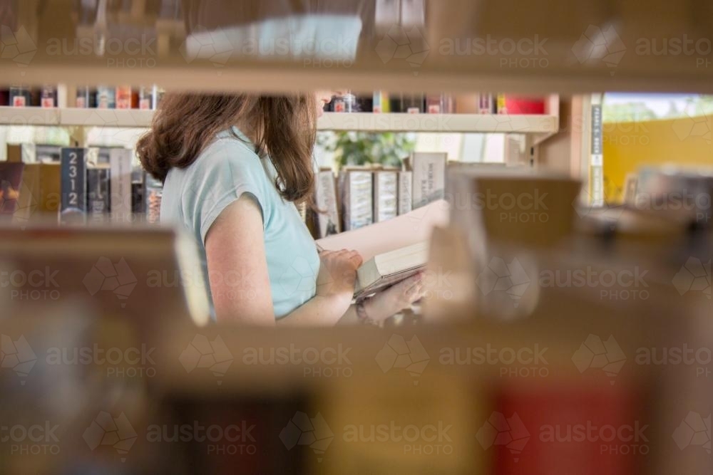 Looking through shelves at a teen girl choosing a book to borrow from the library - Australian Stock Image