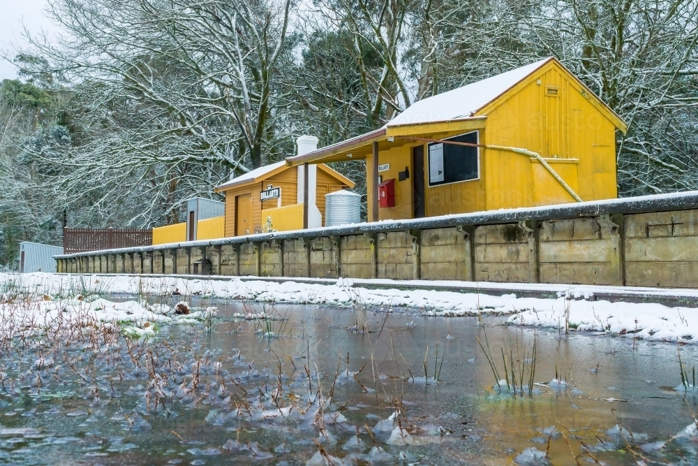 Low angle view of icy puddles in front of a small yellow railway station covered in snow - Australian Stock Image