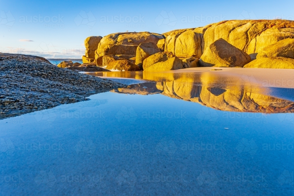 Low angle view of large granite boulders on a beach reflected in a still rock pool - Australian Stock Image