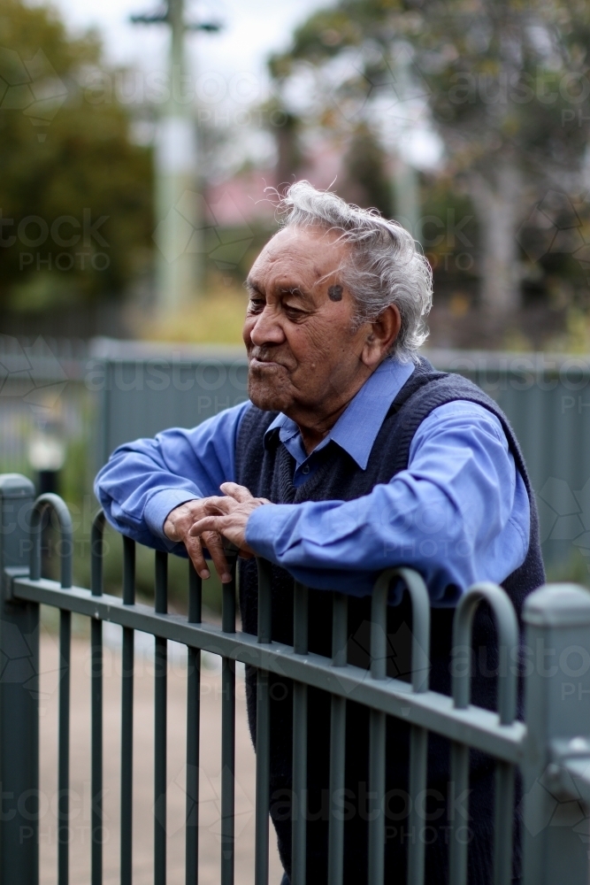 Male Aboriginal elder leaning against fence looking away from camera - Australian Stock Image