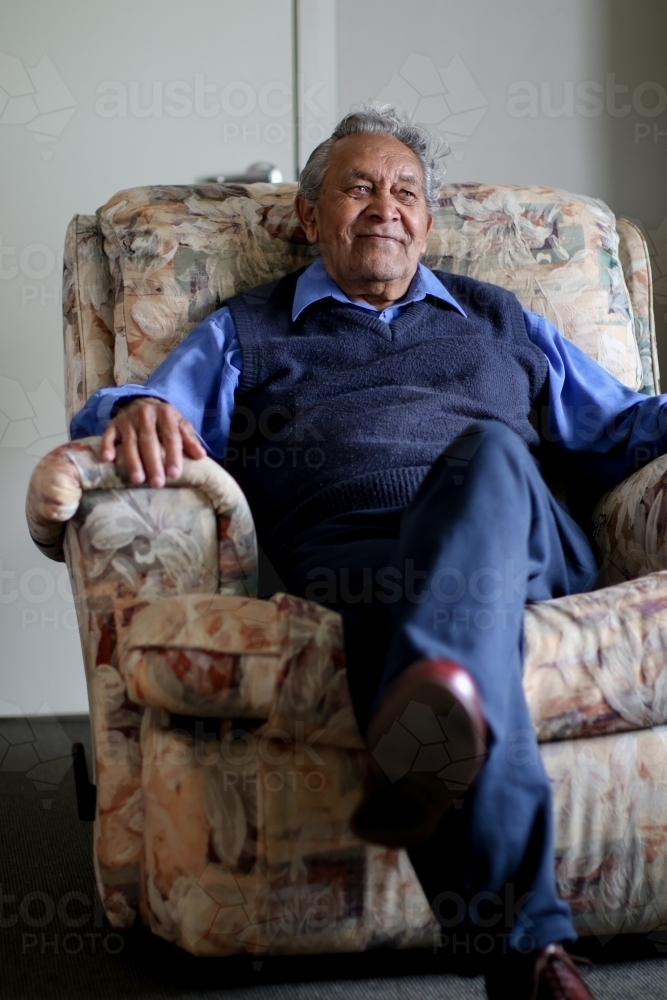 Male Aboriginal elder sitting in a lounge chair in his home laughing - Australian Stock Image