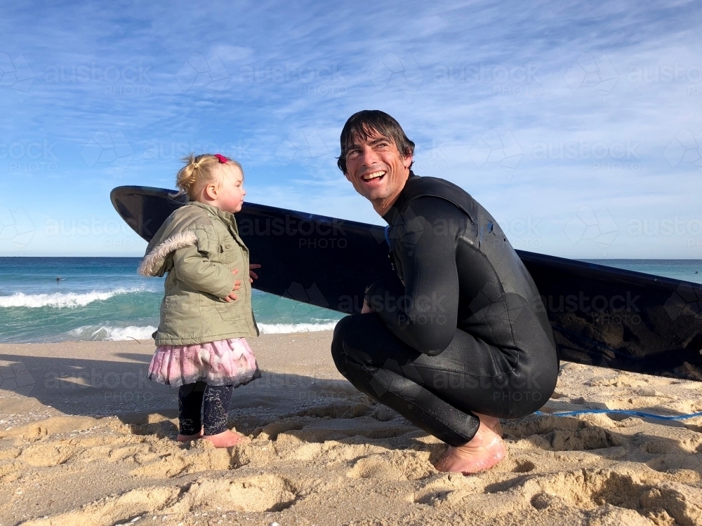 Male adult who’s just got out of surf kneeling down with female toddler on shoreline of beach - Australian Stock Image