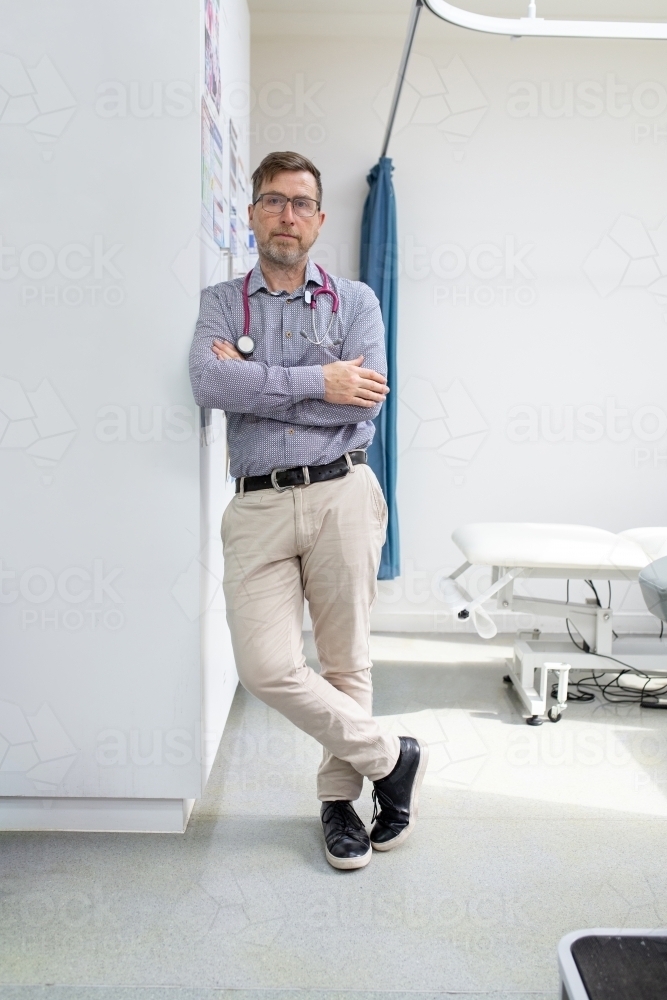 Male doctor with stethoscope around his neck standing while leaning on the wall in the clinic - Australian Stock Image