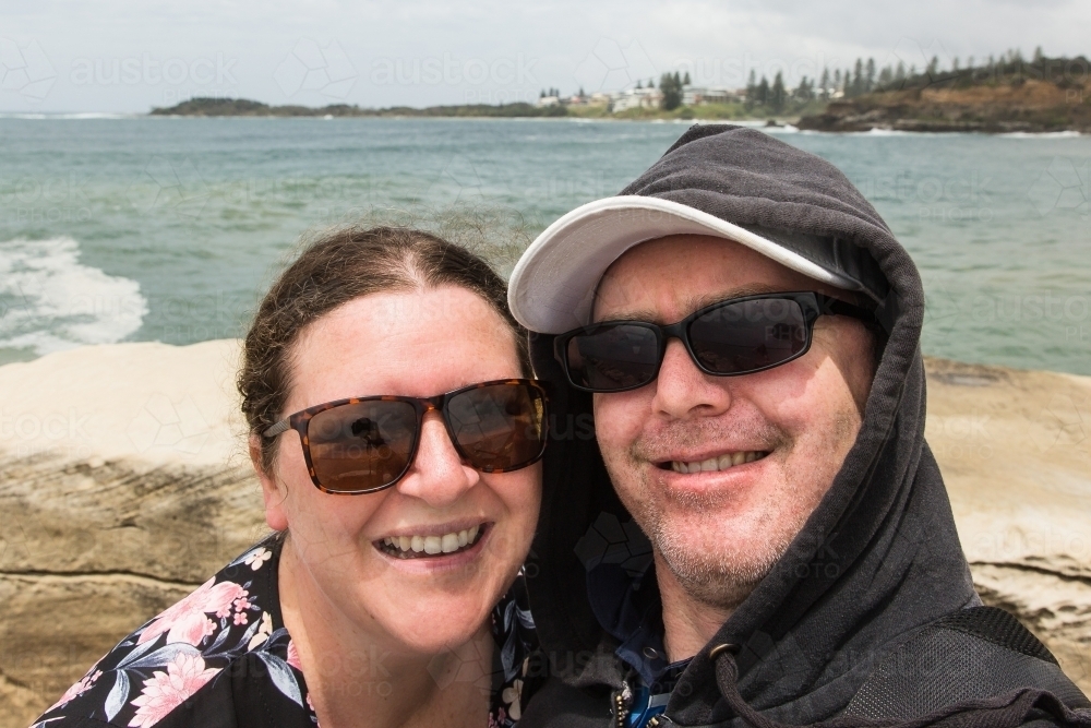 Man and woman selfie - husband and wife smiling wearing sunglasses at beach with ocean behind - Australian Stock Image