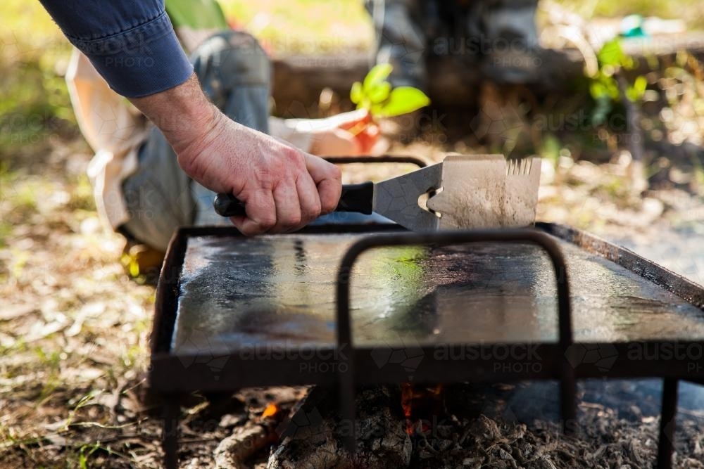 Man cleaning hot plate for cooking sausages on campfire - Australian Stock Image